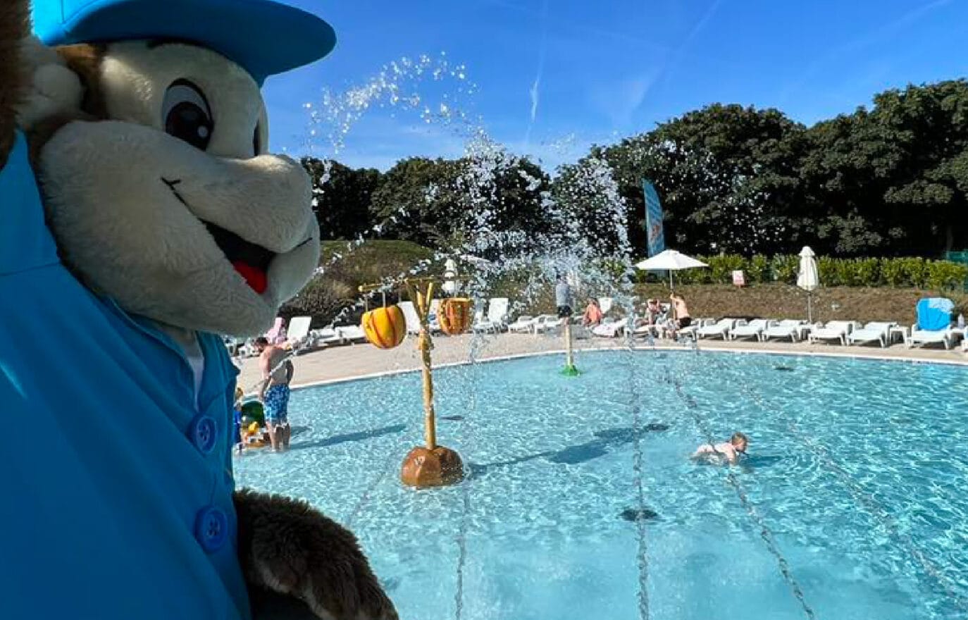 A mascot dressed as a cheerful bear in a blue shirt overlooks a bustling outdoor swimming pool with people enjoying the water and sun loungers around under a clear blue sky.