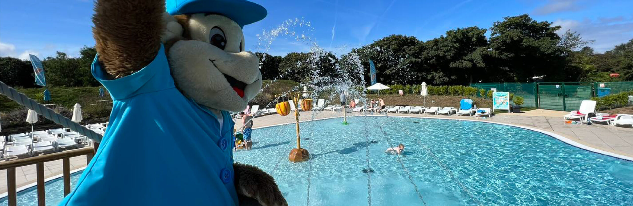 A mascot dressed as a cheerful bear in a blue shirt overlooks a bustling outdoor swimming pool with people enjoying the water and sun loungers around under a clear blue sky.