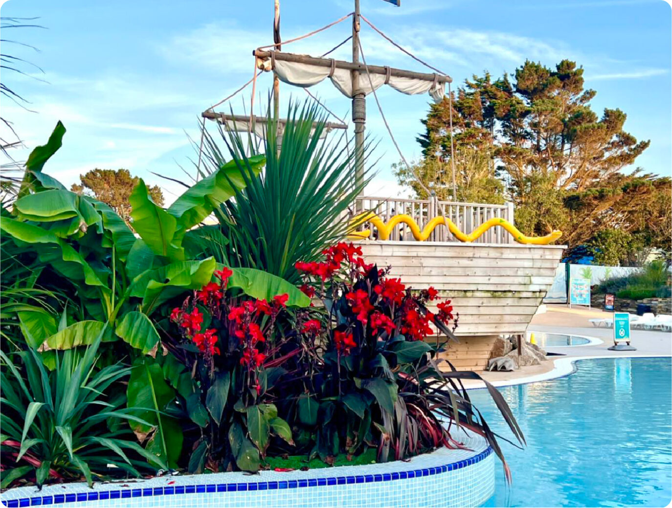 A vibrant outdoor swimming pool scene with lush green plants and red flowers in the foreground, and a whimsical small white boat with a sail structure by the poolside under a clear sky.