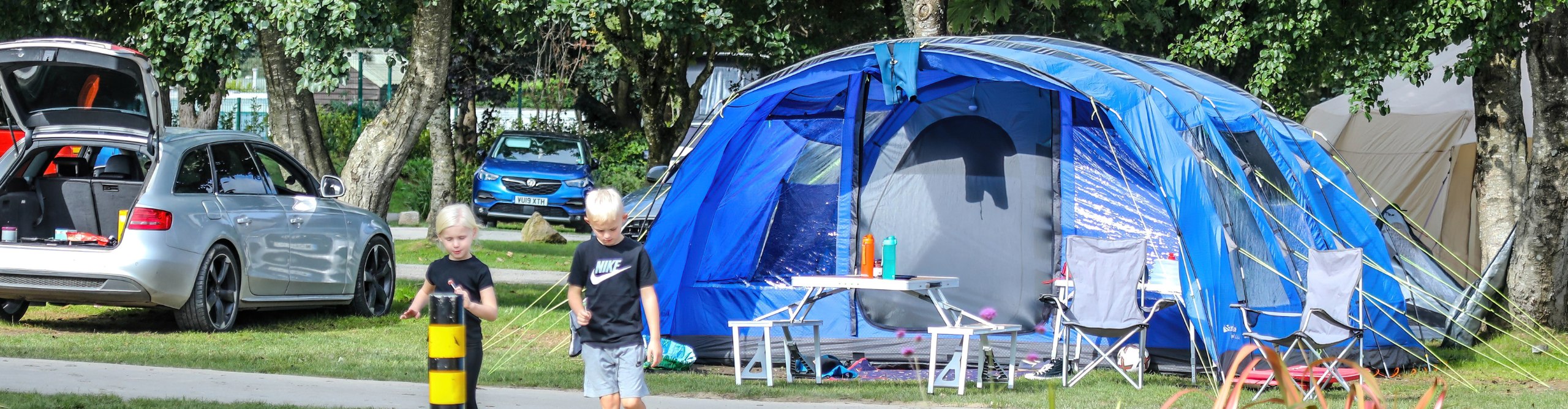 Two boys walking near a large blue camping tent with an open front, next to a parked car in a grassy camping area at Monkey Tree Holiday Park in Cornwall, with trees and another car in the