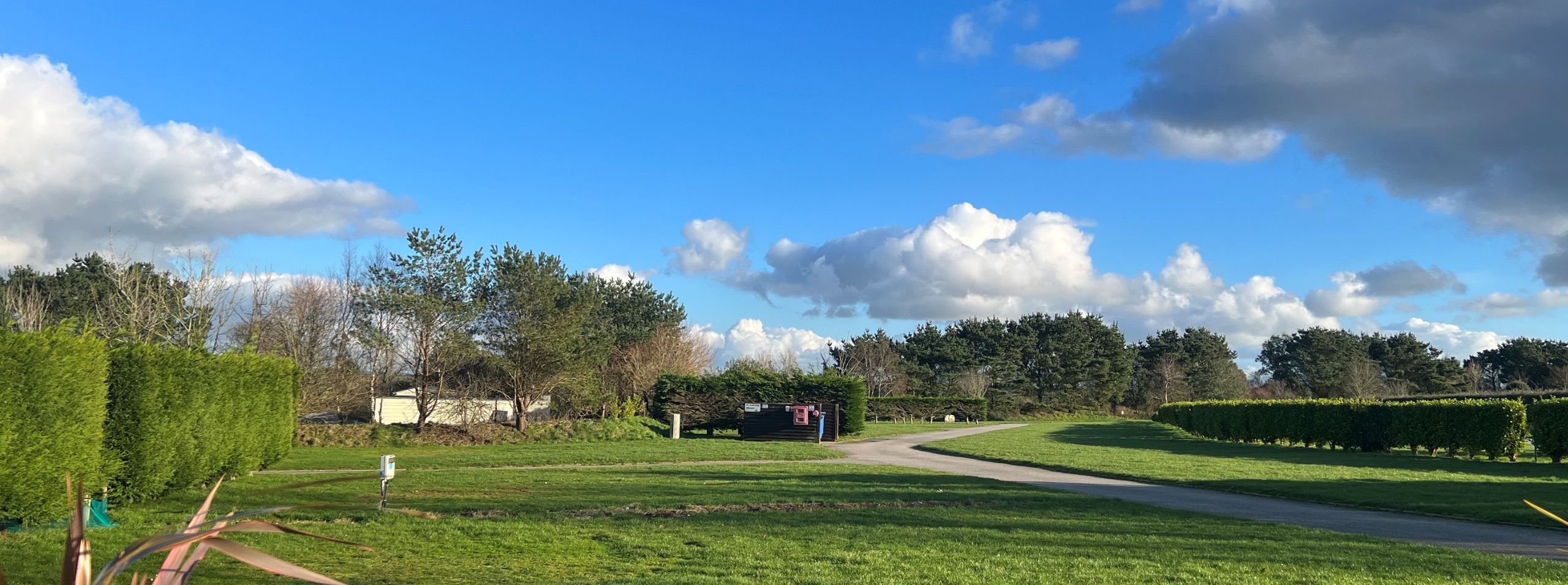 A panoramic view of a lush green park with neatly trimmed hedges, a winding pathway, and fluffy clouds in a blue sky. A small red structure is visible in the distance at the Cornwall campsite