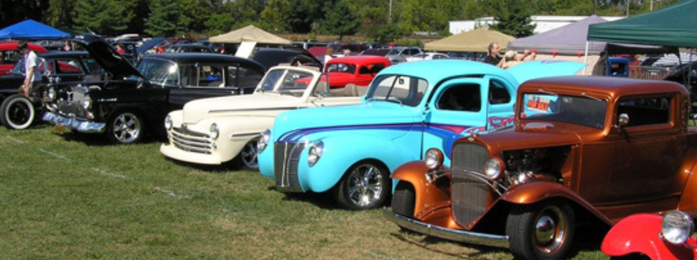 Outdoor car show featuring a row of vintage cars in a field, with models varying in color and style, under clear skies. People admire the vehicles in a leisurely luxury setting.