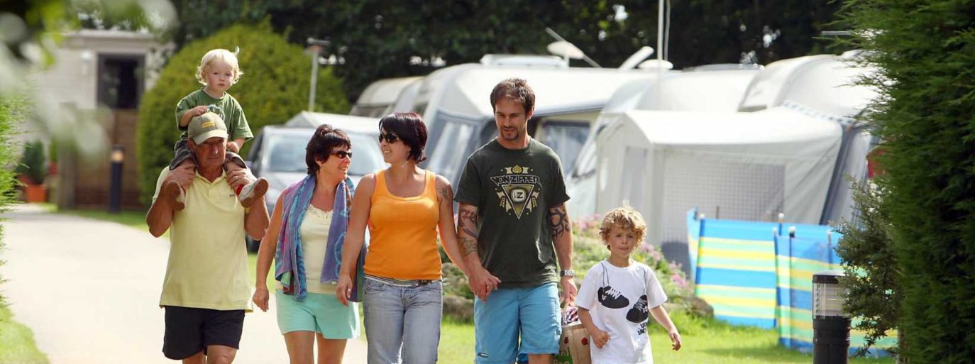 A family enjoys a sunny day at a campground in Cornwall, with two adults carrying young children and a third child walking alongside, surrounded by parked trailers and lush greenery.