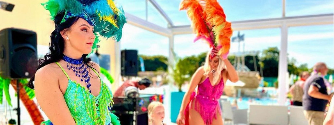 Two women wearing vibrant carnival costumes with feathered headdresses perform at a luxury poolside event under a clear blue sky.