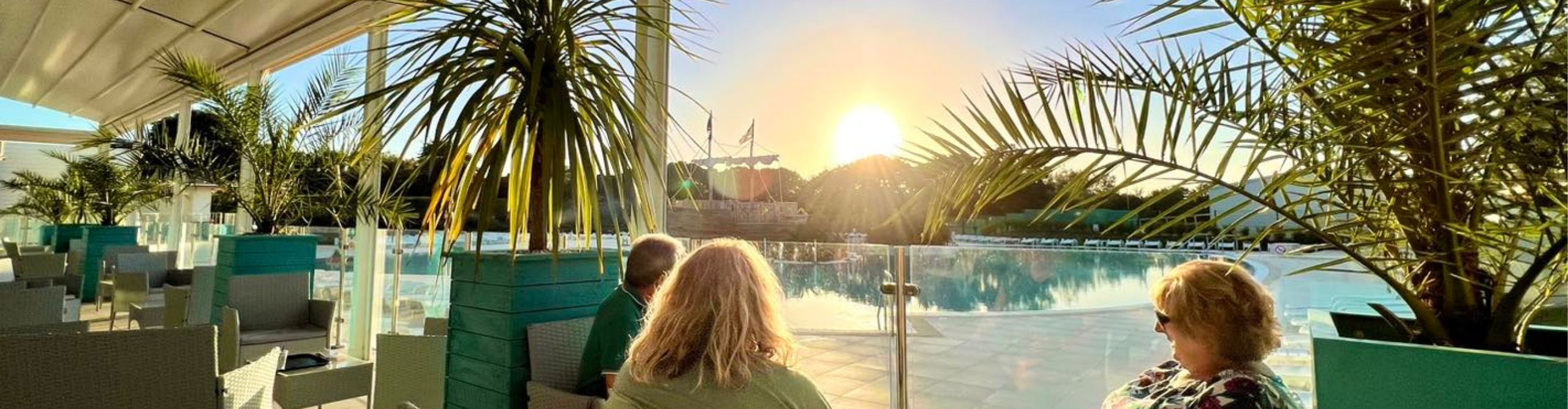 Two people sitting and enjoying a sunset view over a pool surrounded by palm trees, with a picturesque pirate ship in the background at Monkey Tree Holiday Park in Cornwall.