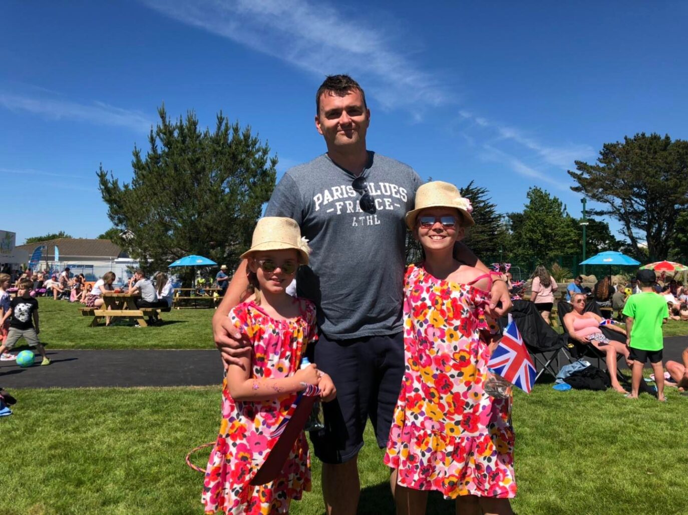 A man stands outdoors with two young girls wearing matching floral dresses and straw hats. They smile in a sunny park filled with people, and British flags are visible in the background, celebrating a holiday near Cornwall