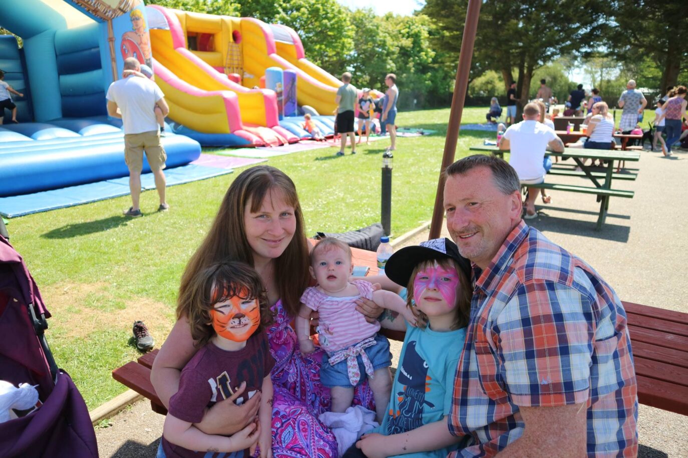 A family enjoying a sunny day at a park with a luxury bouncy castle in the background. The children have face paint, one as a tiger, and they are all smiling. There are other people
