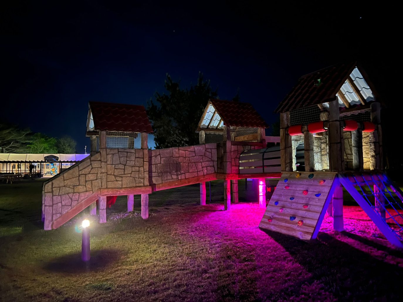 A nighttime image of an illuminated playground featuring a climbing wall, slides, and monkey tree, highlighted with vibrant purple and red lights under a starry sky.