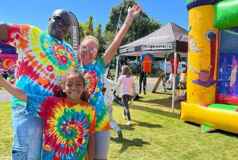 Three people wearing colourful tie-dye shirts smile and pose with arms outstretched at an outdoor event. There is a bouncy castle, a tent labelled surf lessons, and sunny weather in the background.