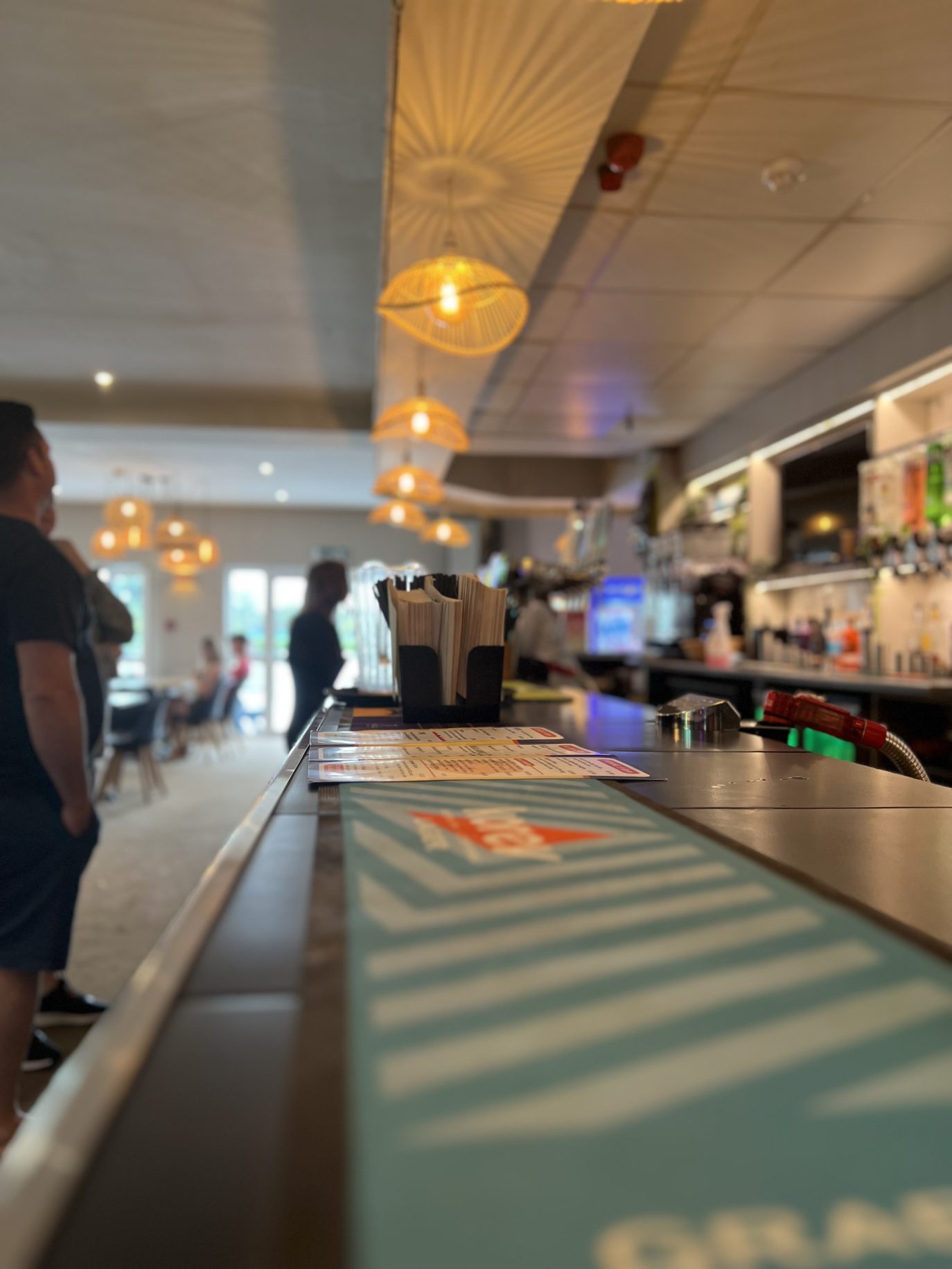 Blurred interior of a luxury café with sharp focus on a blue menu on a counter, overhead pendant lights, and customers standing and sitting in the background.