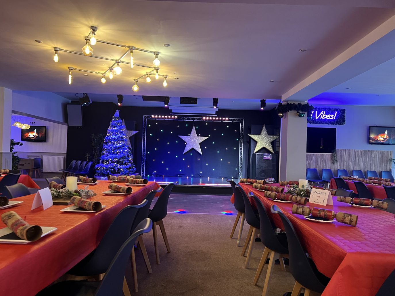 A festive event hall with a stage decorated with star motifs and blue lighting, surrounded by tables set with red tablecloths, christmas crackers, and a large, lit monkey tree to the left.