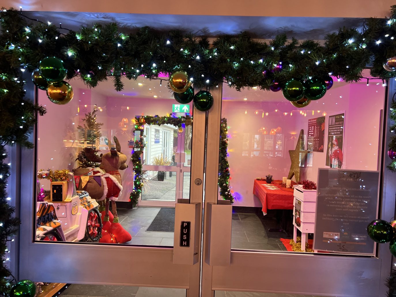 A festive store entrance decorated with Christmas garlands and baubles. Inside, a brightly lit space features a Christmas tree, reindeer statues, and a table covered with gifts at the Monkey Tree Holiday