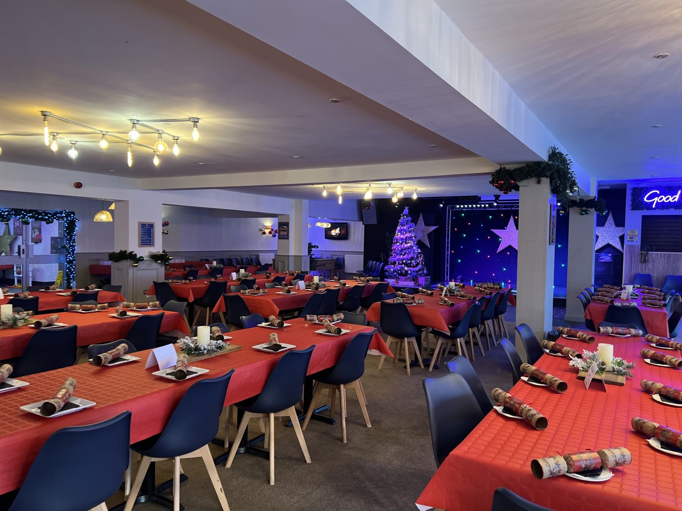 A festive banquet hall decorated for Christmas with red tablecloths, set tables, and a lit Christmas tree next to a stage with a 
