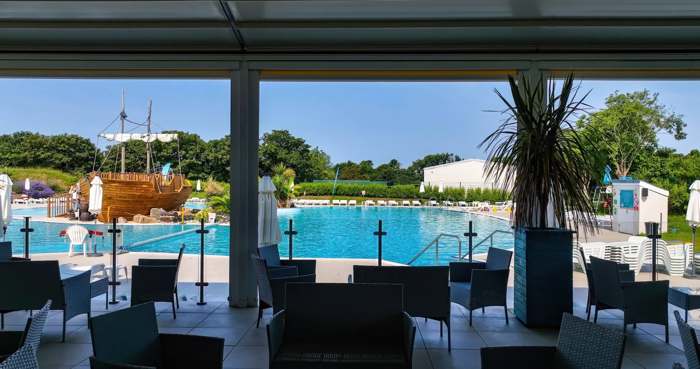 View from a shaded area with chairs and tables overlooking a vibrant outdoor pool with crystal blue water and a decorative wooden ship, situated in Cornwall’s lush green landscape under a clear sky.