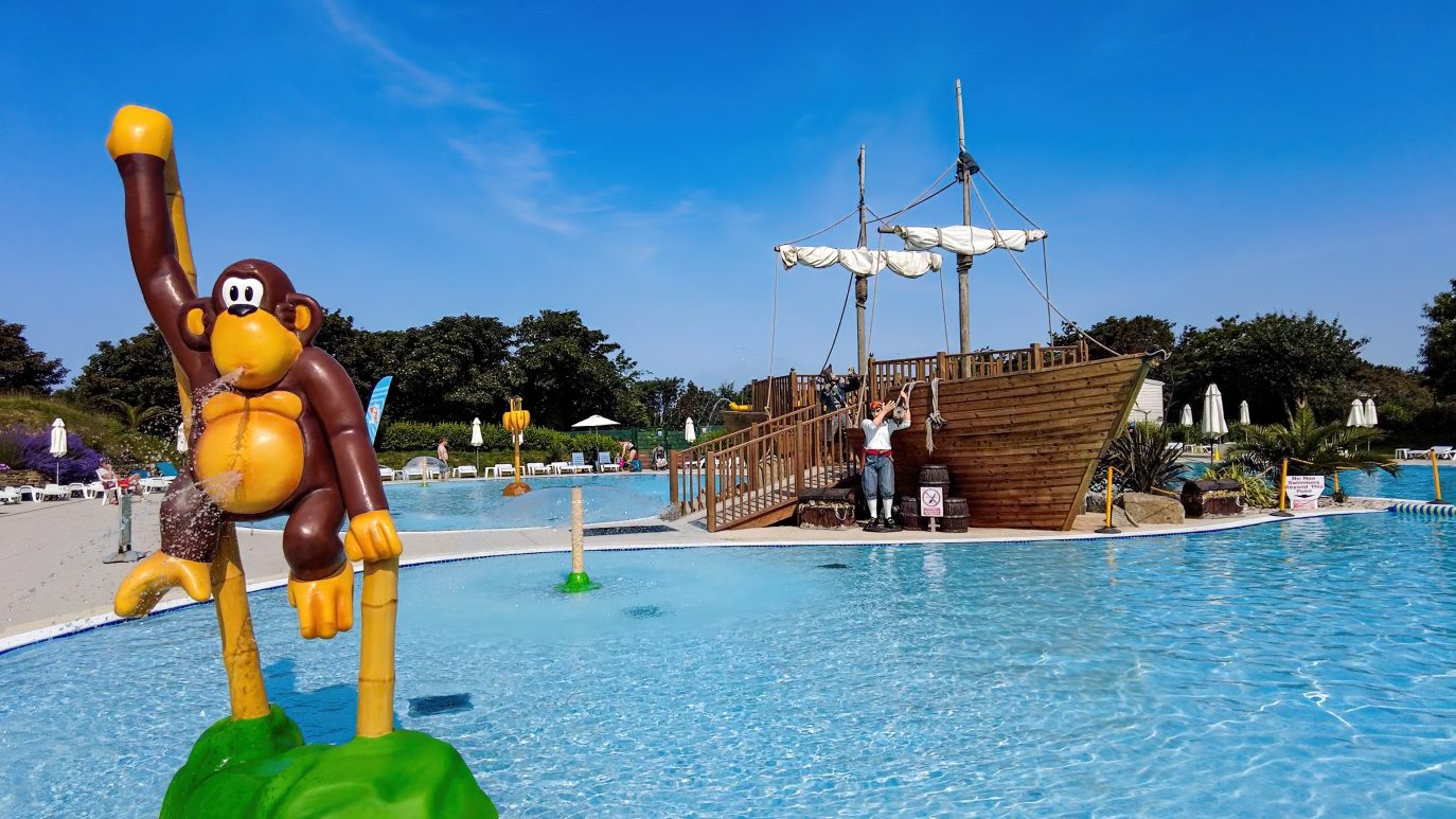 A large inflatable monkey holding a banana stands in a shallow pool at a water park, with a wooden pirate ship replica and visitors enjoying the sunny day in Cornwall.