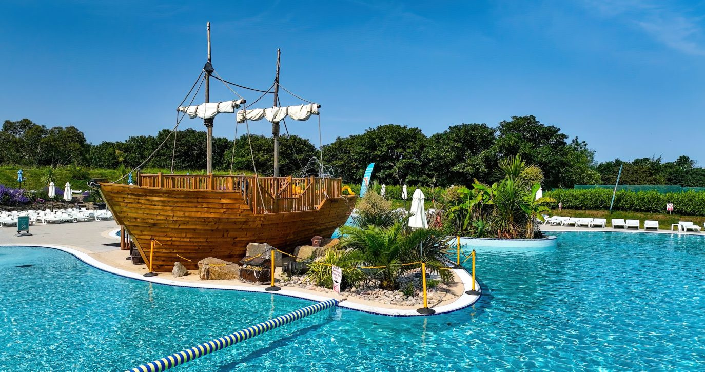 A wooden pirate ship with sails down, floating on a calm, bright blue pool surrounded by lush greenery and clear skies at a holiday home in Cornwall.