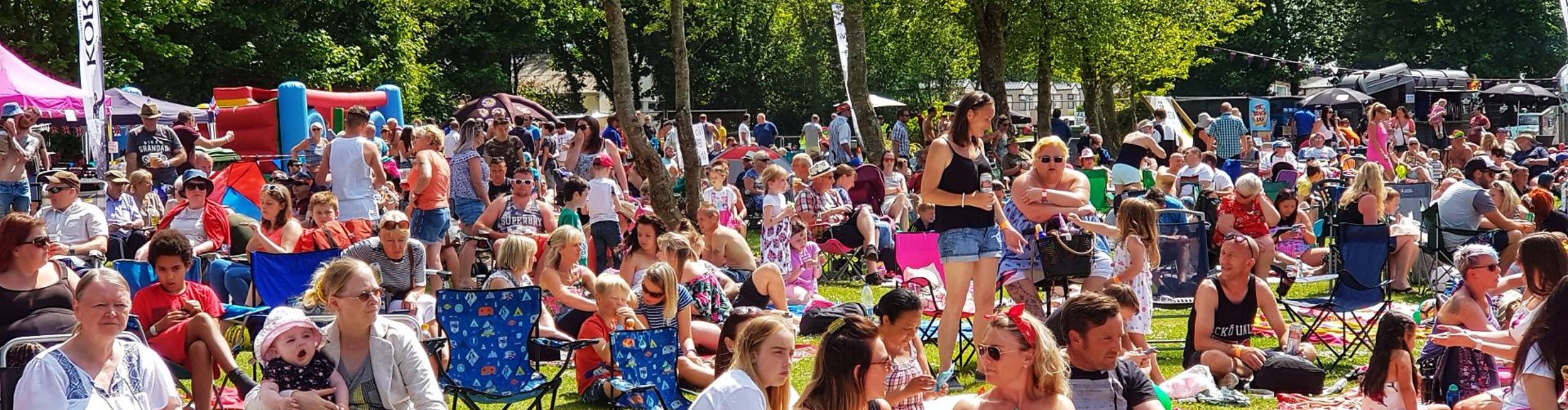 A vibrant outdoor event in Cornwall with a diverse crowd of people seated and standing on a grassy area, enjoying a sunny day. Some are holding umbrellas, and children are visible. There are tents