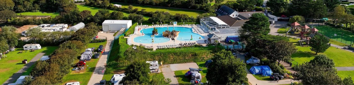 Aerial view of a vibrant holiday campsite featuring a large swimming pool, parked RVs, and green spaces, surrounded by trees under clear blue skies.