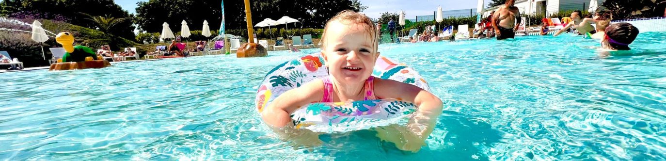 A toddler with a joyful expression, wearing a colorful float ring, wades in a sunny outdoor pool at a holiday home in Cornwall, surrounded by other swimmers.