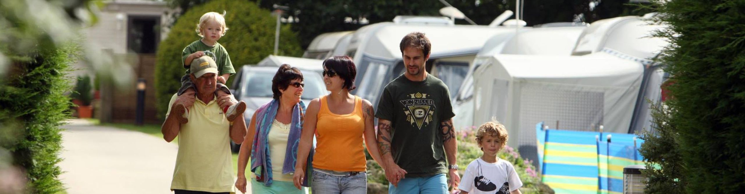 A multigenerational family walking and chatting on a sunny day at a campground in Cornwall, with trees and holiday homes in the background. An older man carries a young child on his shoulders.