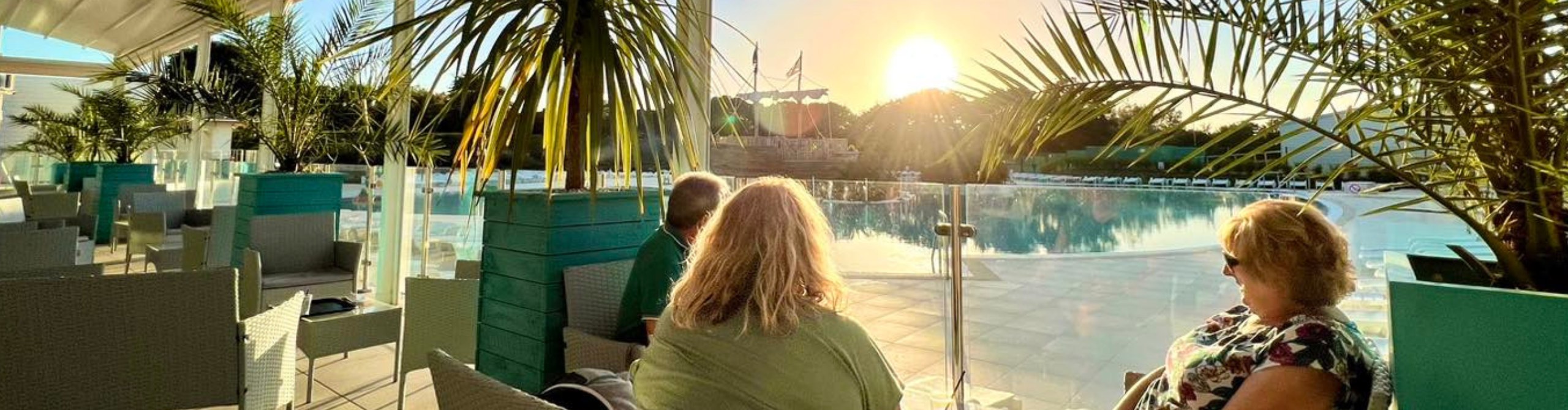 Three people relaxing on patio chairs under palm fronds by a tranquil poolside at sunset, with a soft golden light casting long shadows in Cornwall.