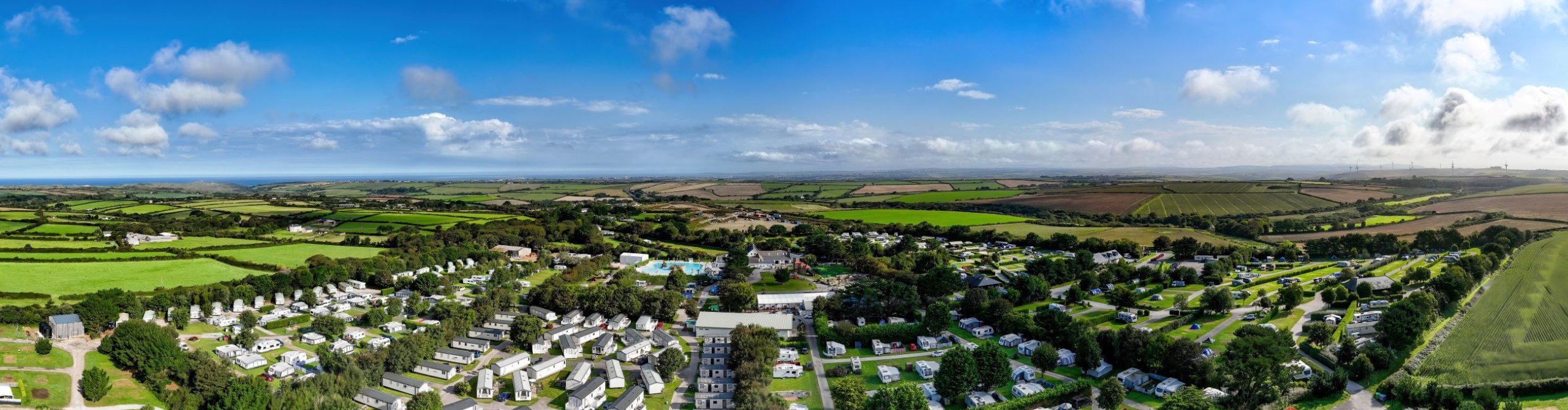 Panoramic aerial view of a lush rural Cornwall landscape featuring a large campground with numerous holiday homes, surrounded by expansive green fields and a clear blue sky.