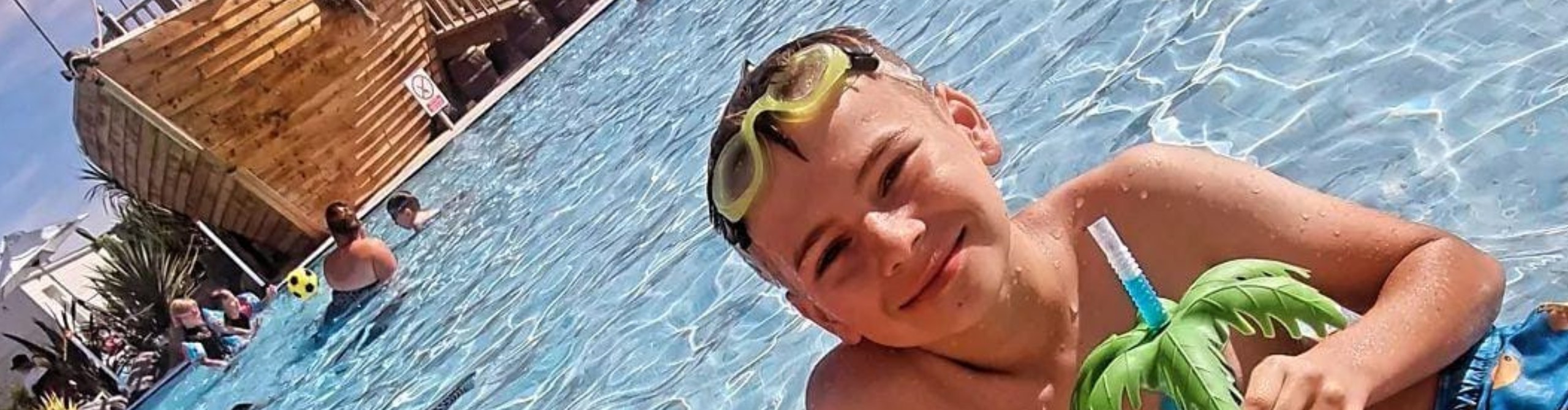 A young boy wearing goggles and a swimming tube, smiling broadly in a sunlit pool at a holiday home with other swimmers and a wooden structure in the background.