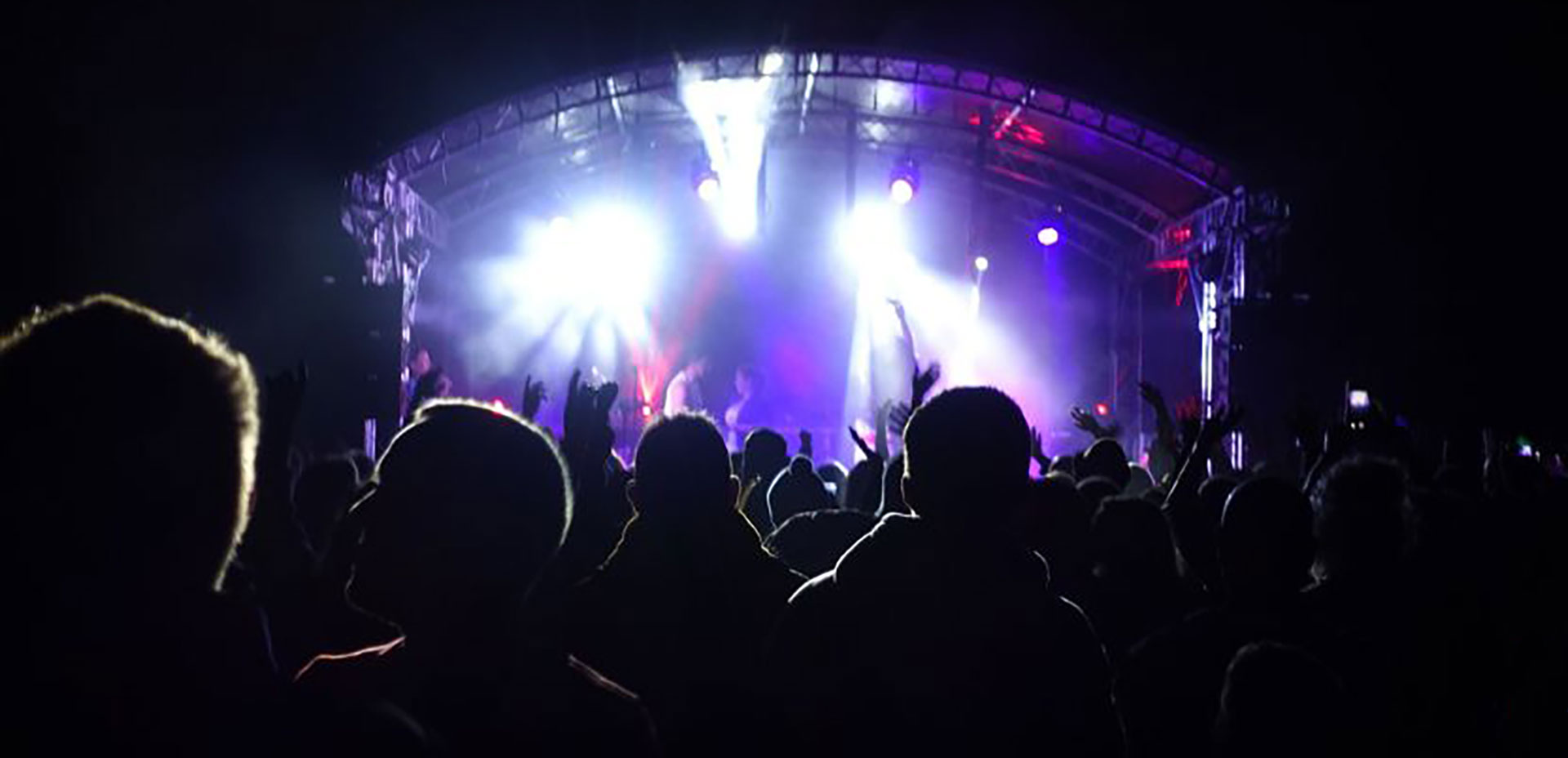 A backlit view of a crowd at a concert, facing a brightly lit stage with dramatic purple and blue lights illuminating performing artists during a camping festival.