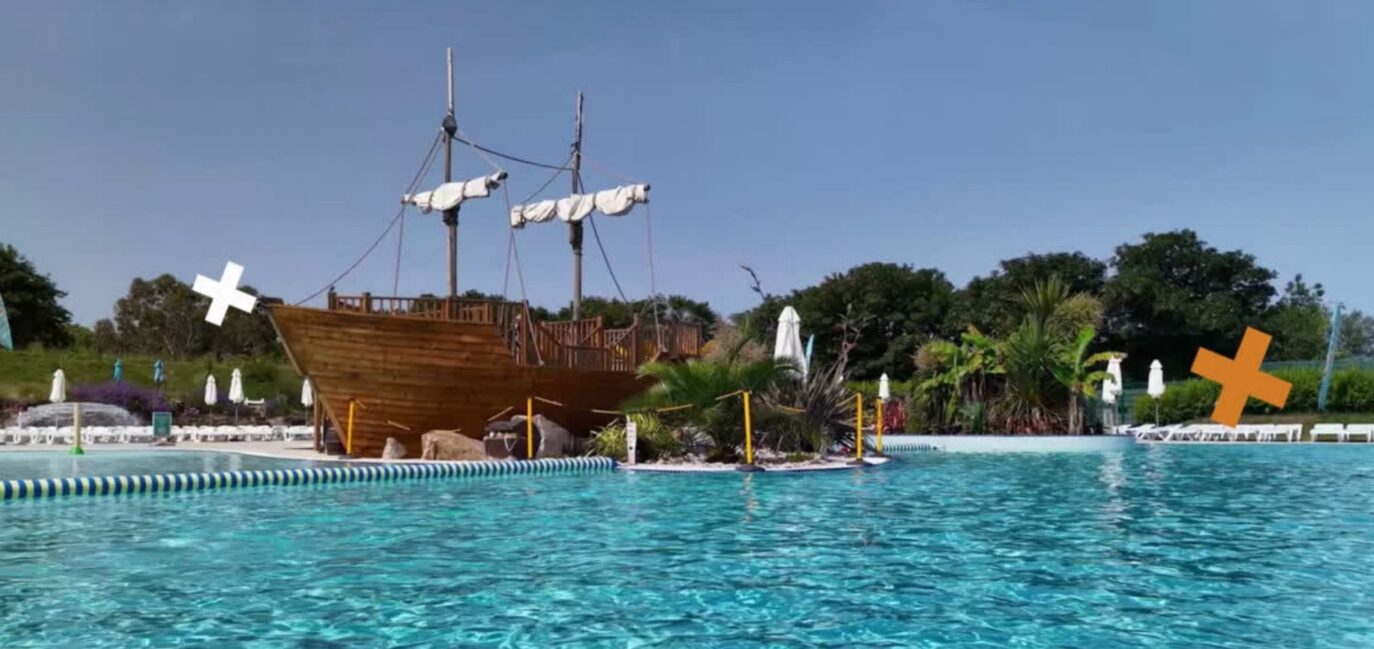 A large pirate ship replica emerges from a blue swimming pool surrounded by lounge chairs and lush monkey trees under a clear sky.