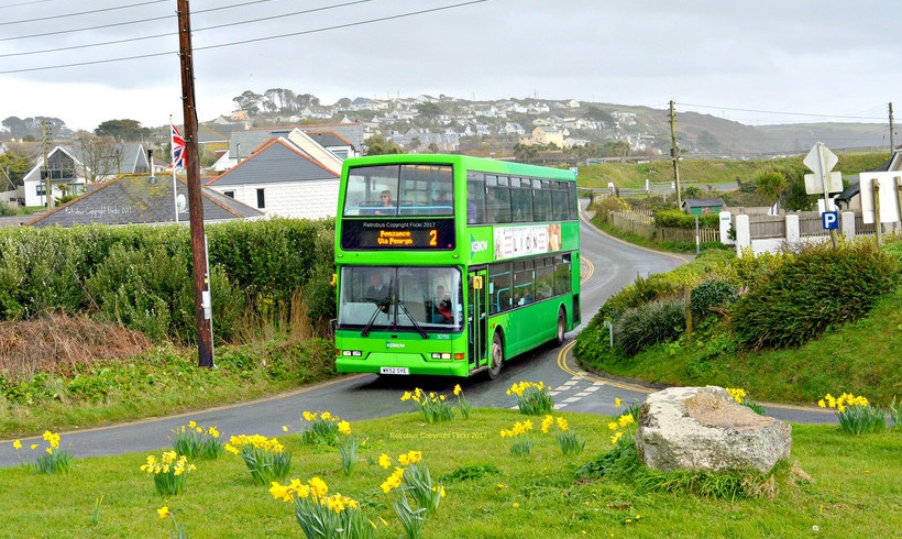 A green double-decker bus offering bus services from Monkey Tree Holiday Park winds along a rural road lined with yellow daffodils, passing houses and hills under a cloudy sky.