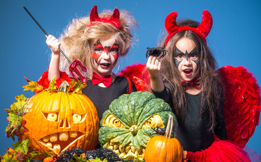 Two children in red devil costumes with face paint pose excitedly with Hallowe’en pumpkins, one holding a fake spider. Surrounded by autumn leaves against a blue background, they capture the playful spirit of Halloween Cornwall.