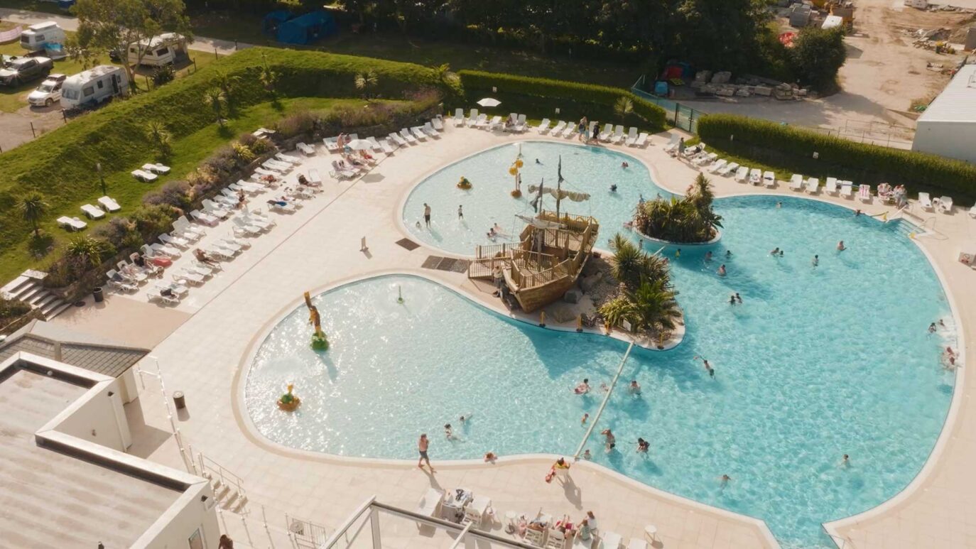 Aerial view of a large outdoor pool shaped like a heart, featuring a pirate ship structure in the middle—a perfect spot for Cornish holidays. People swim and lounge on chairs around the pool, bordered by lush green hedges and trees, creating a tranquil camping escape.