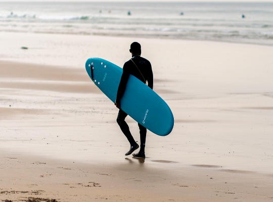 A man holding a surfboard on one of the best beaches near Newquay, Cornwall, with golden sand and clear blue water in the background.