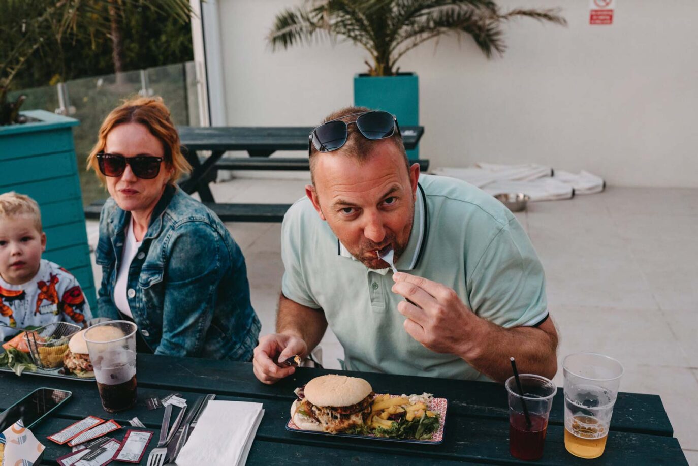 Family dining outdoors at Monkey Tree Holiday Park, with a man enjoying a burger and fries and a child seated nearby.