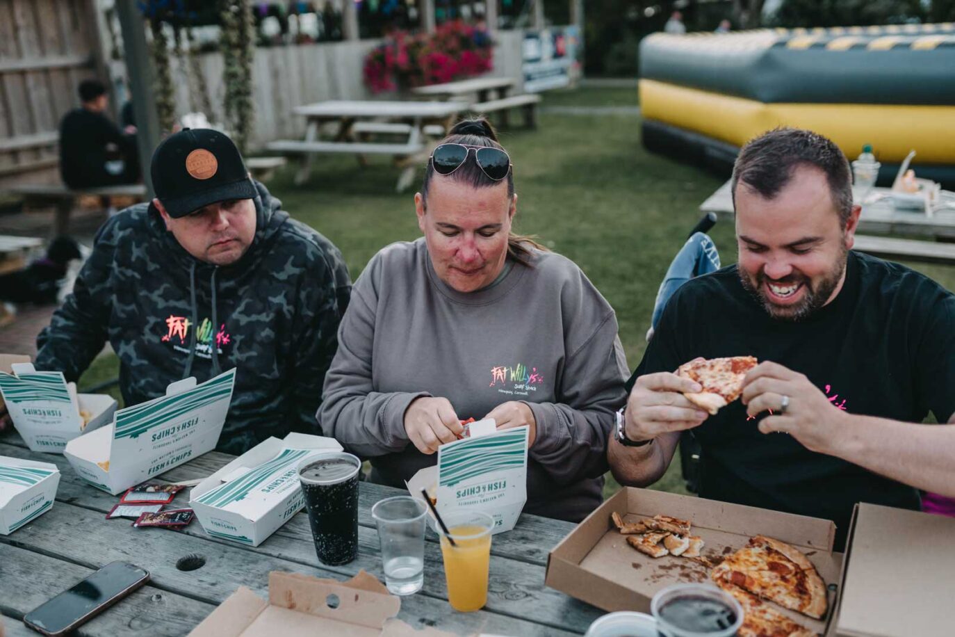 Adults smiling and eating fish and chips and pizza at a picnic table in the food court area at Monkey Tree Holiday Park.