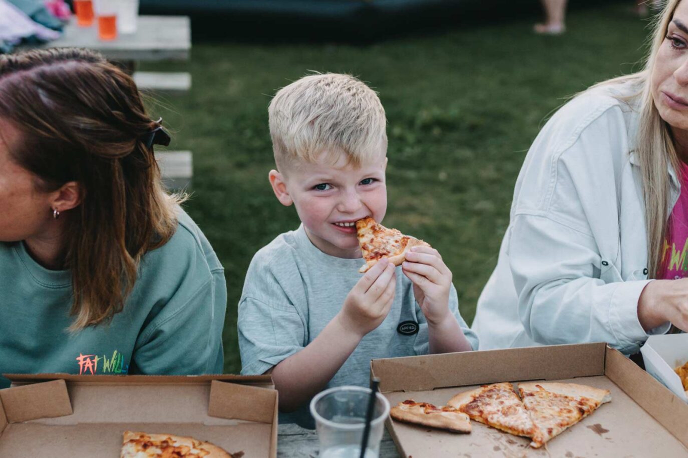 Young boy smiling while eating pizza at a picnic table, surrounded by family during a relaxed meal at Monkey Tree Holiday Park.