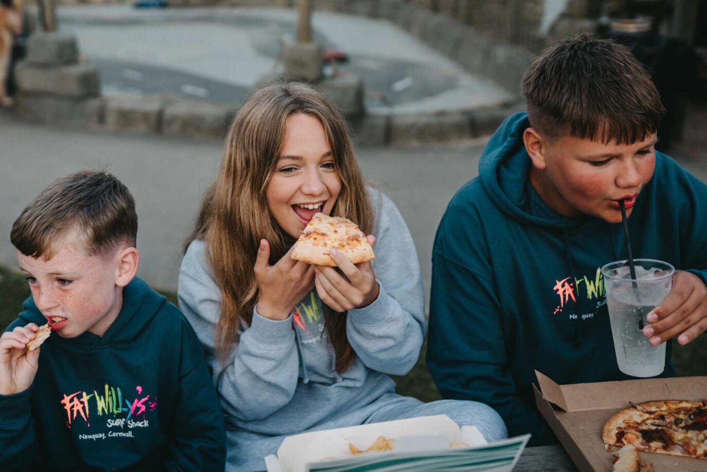 Three children enjoying pizza and drinks outdoors at Monkey Tree Holiday Park, wearing Fat Willy’s hoodies.