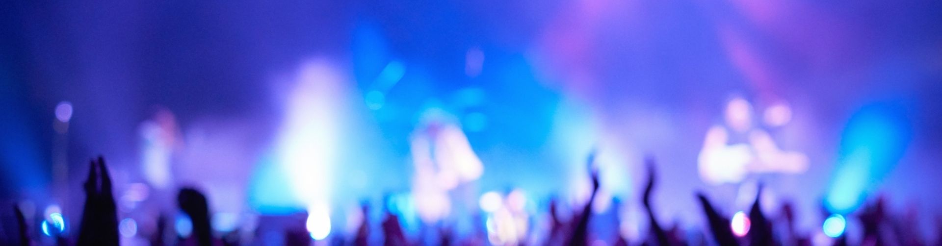 Blurry image of a concert during the October festival weekend, with bright purple and blue stage lights, a crowd of people raising their hands, and band members performing on stage in the background.