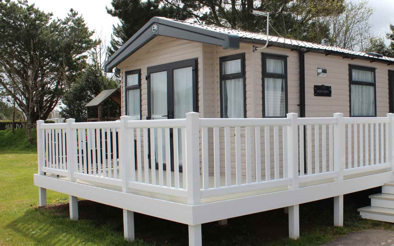 A modern mobile home with large windows and a white railed decking sits elevated on a grassy area, surrounded by trees and greenery on a sunny October festival weekend.